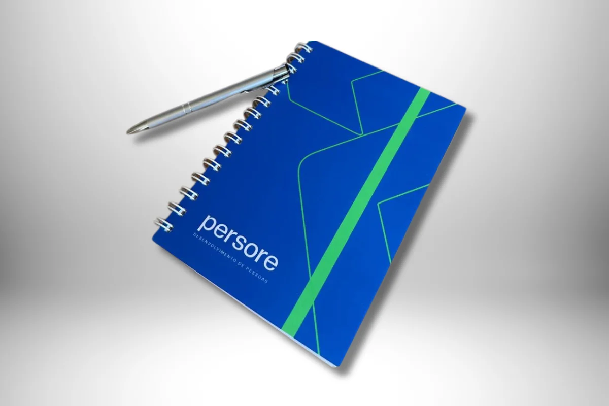 Agenda personalizada espiral da marca Persore com capa azul e design corporativo, acompanhada de uma caneta metálica.